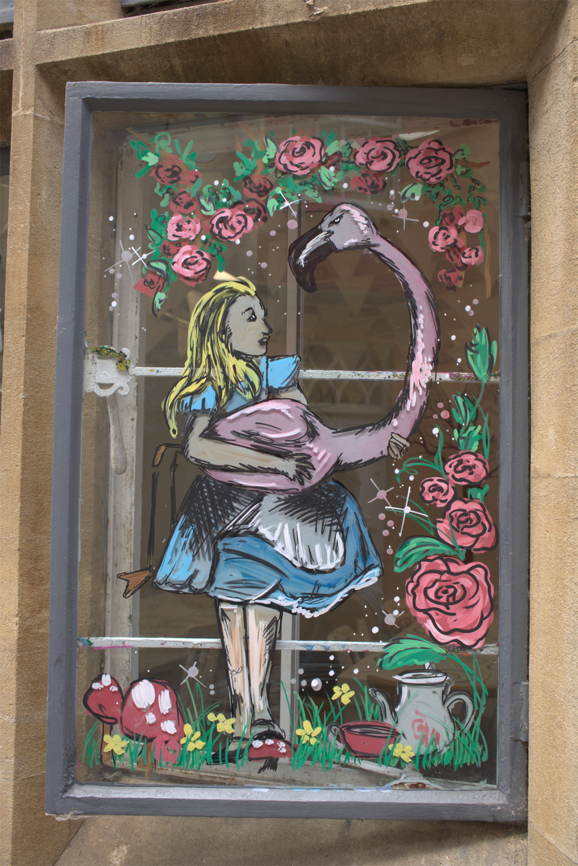 Alice in Wonderland mural drawn with markers on a glass window. Alice stands on a field of flowers and mushrooms, wearing her well-known knee-length blue puffed dress with a white pinafore apron. She is holding a stern eyed pink flamingo whose neck arcs up over Alice's head. Alice and the flamingo are surrounded by a frame of roses. The window frames the mural and contrasts with a black frame. (RAW)