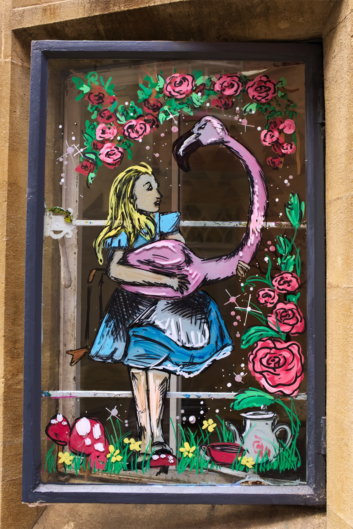 Alice in Wonderland mural drawn with markers on a glass window. Alice stands on a field of flowers and mushrooms, wearing her well-known knee-length blue puffed dress with a white pinafore apron. She is holding a stern eyed pink flamingo whose neck arcs up over Alice's head. Alice and the flamingo are surrounded by a frame of roses. The window frames the mural and contrasts with a black frame. (Edit)